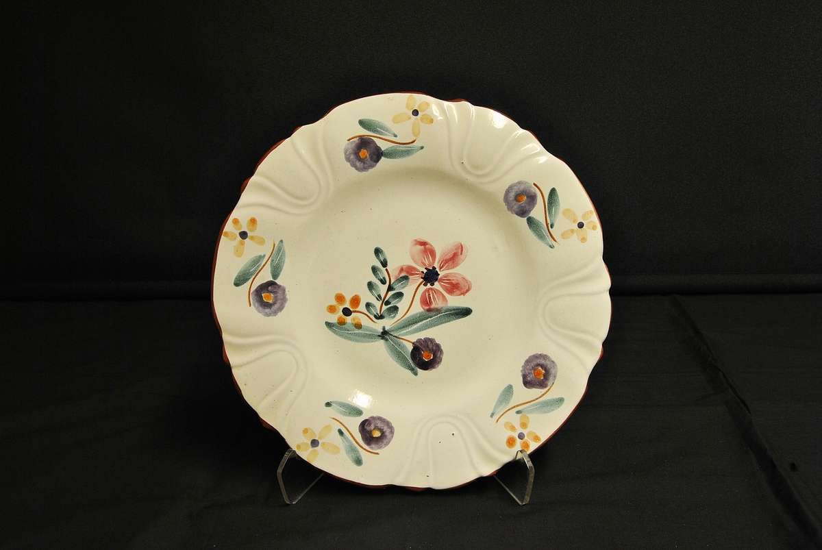 Sirkelforma, kvit tallerken med bølga kant og blomsterdekor: eit stort motiv på midten og fem mindre motiv rundt kanten.