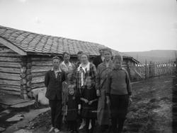 Gruppe barn og voksene utenfor en seterbygning i tømmer. Bla