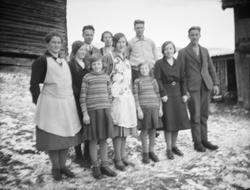 Voksne og barn oppstilt ved tømmervegg med snø på bakken.