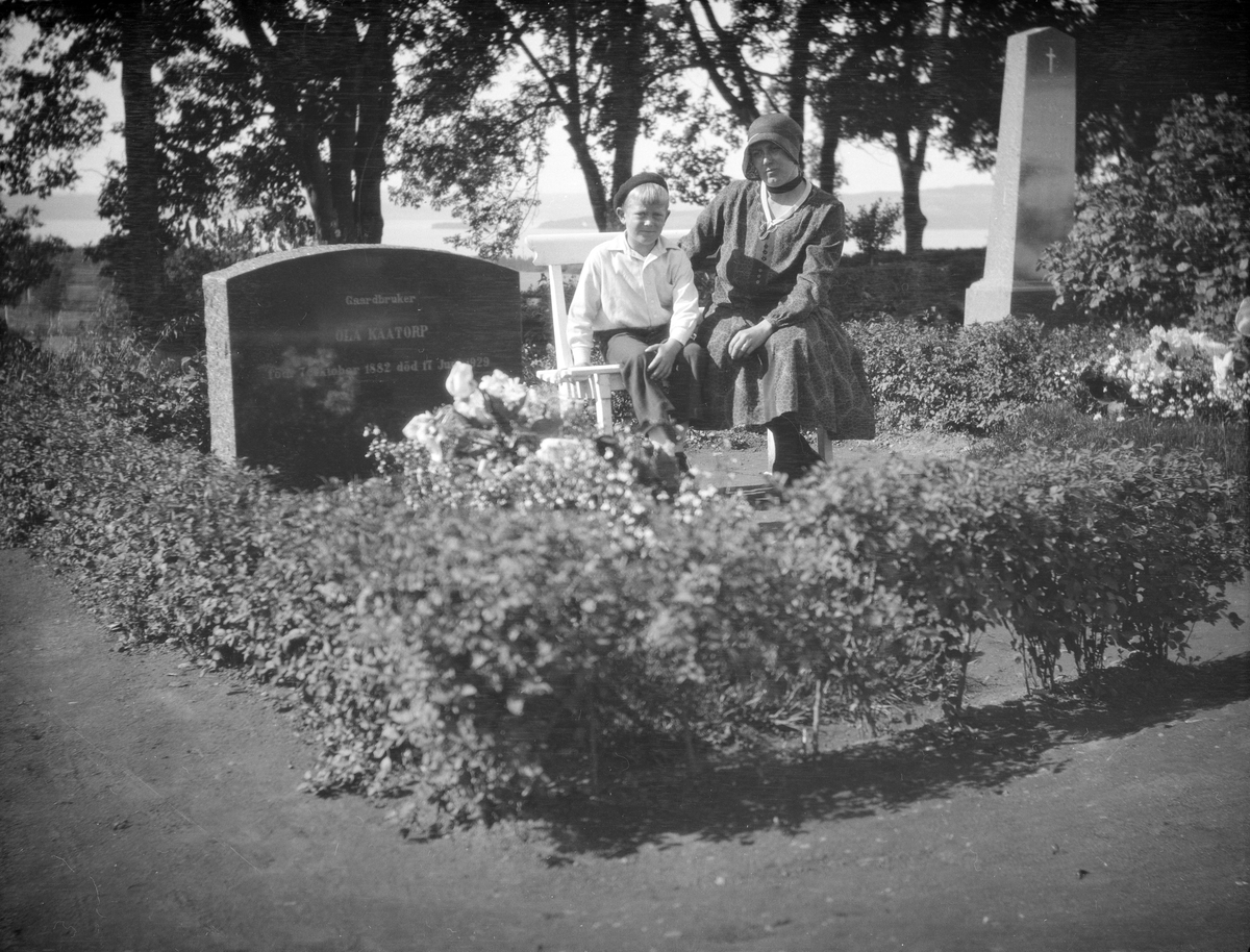 Kvinne og gutt sitter på en benk ved gravsteinen til Olav Kaatorp f. 1882 som ligger på Furnes kirkegård.