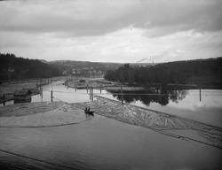 Stor elv med tømmerlenser hvor to menn er i båt og i arbeid.
