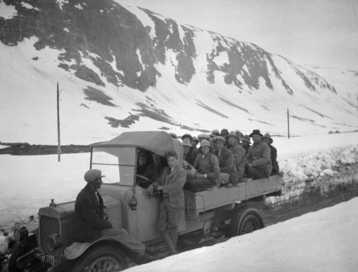 En lastebil full med folk på lasteplanen står på veg mellom brøytekanter på Strynsfjellet. Lastebilen E-1574 i fra Oppland fylkesveivesen, Staværn bilfabrik A/S 1921 modell, chassisnummer: 21334