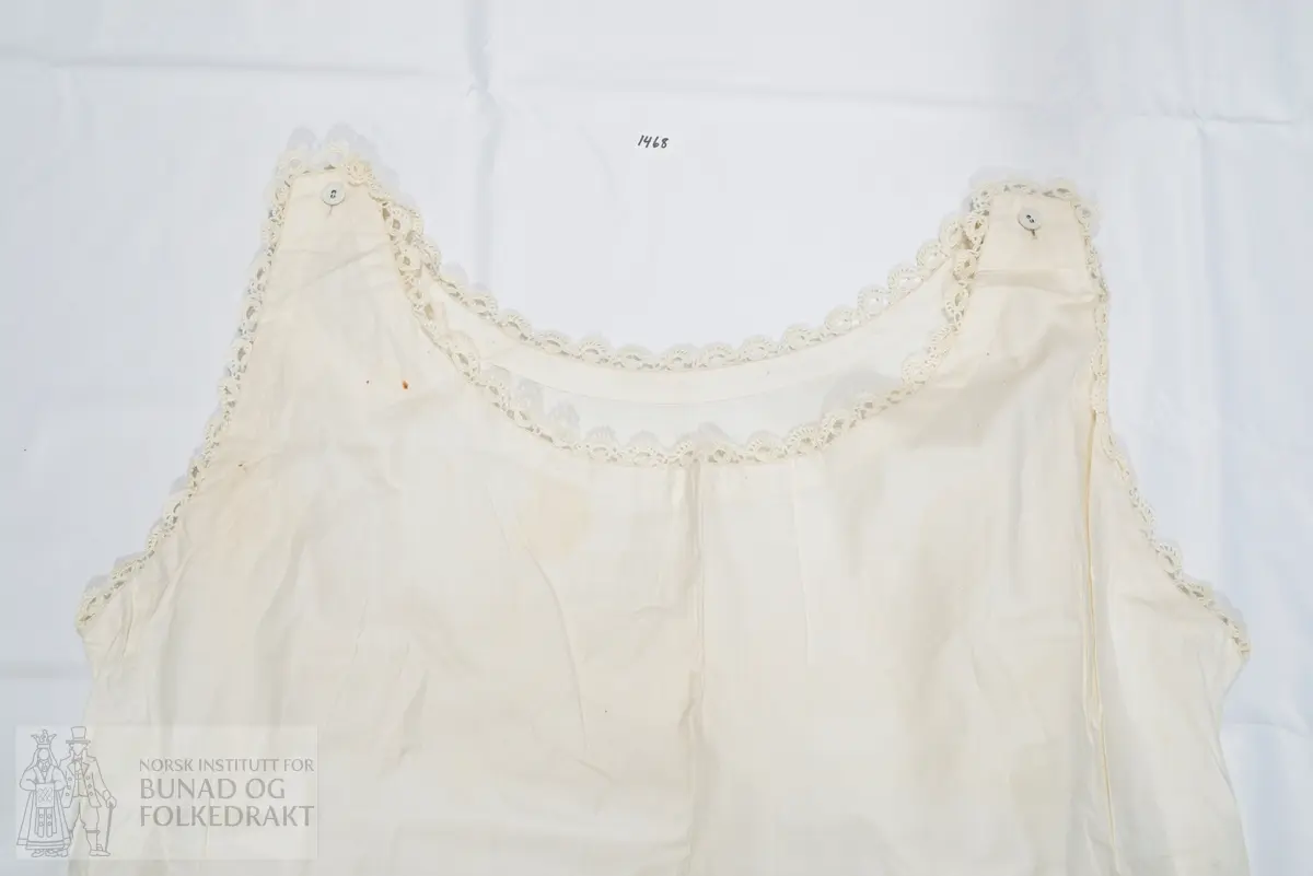 Nattkjole i bomullslerret med smal heklet kant rundt hals og ermringing. Sydd av et for- og bakstykke, med knepping med knapphull og knapp på skuldrene. Sydd sammen med indresøm på maskin. Svært små sting. Den påsydde blonden er heklet av mercerisert bomullsgarn. Dobbel oppbrett nederst på skjørtekanten.