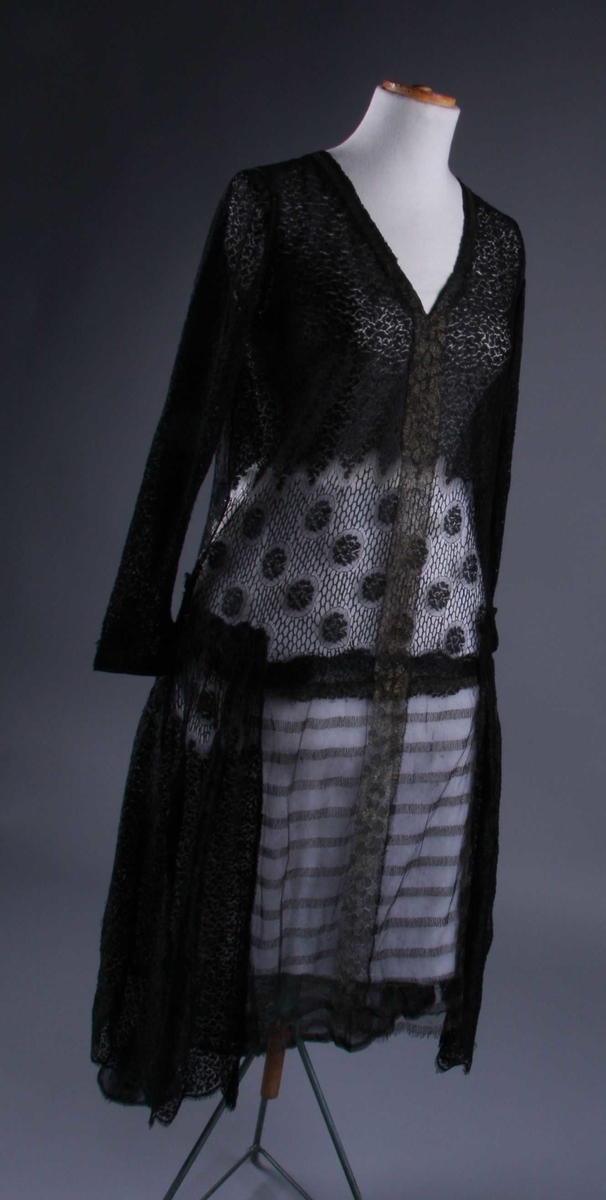 Av sort silkeblonde og metall-innvirket tyll. Kjolen har sidt liv med dyp, spiss utringing foran og en mindre bak, begeg med innsatt stykke av metall-innvirket tyll. Skjørtet har for- og bakstykke av samme stoff med tettere bord på midten, og tyllen fortsetter oppover livet like til halsen. Bakstykket er dobbelt; forstykket er kortere fortil enn bak hvor det går i en bue fra sidepanelene. Kjolen har lange, trange ermer med en liten tyll.fichu ved håndleddet. Stort silkebelegg langs kantene, også rundt ermhullet, på retten.