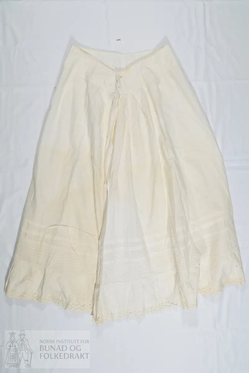 Understakk i hvitt bomullslerret. Bred linning som lukkes med en trukket hvit knapp. Linningen har en skjøt. Flere legg nede på stakkekanten, to bredere og flere mindre. Helt nederst er stakken påsydd en blondekappe, så nederst består stakken av to lag stoff. Blonden er av bomull med engelsk broderi. Stakken er sydd sammen av 5 høyder. Kun maskinsøm.