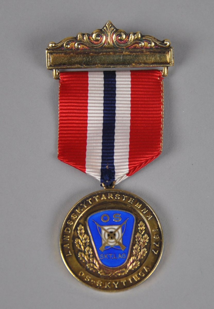 Medalje med logo fra Os skilag. Nål med bånd i rødt, hvit og blått (blåfargen er falmet).