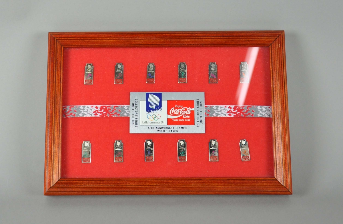 Tolv pins i glass og ramme, med logo for Coca Cola og piktogrammer før ol-øvelsene, på rød bakgrunn. I rammen ligger også et merke med logo for Coca-Cola og Lillehamemr-OL 94.