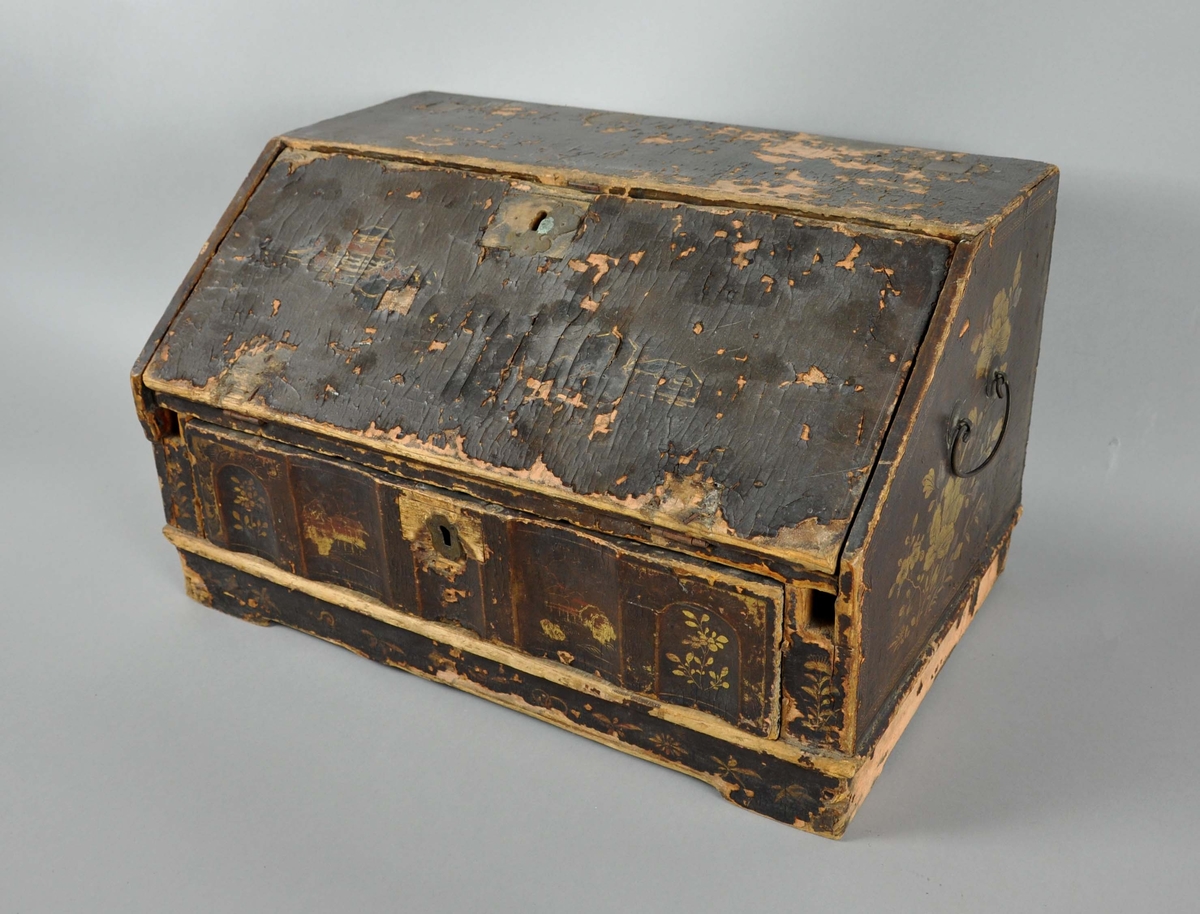 Reiseskrin av tre, rektangulær form med skrå framside. Skrinet har brun grunnmaling og gullfarget dekormaling oppå. Malingen har i stor grad falmet av og bare litt av dekormalingen er synlig. På framsiden av skrinet, nederste del, er det en skuffe med seksjonerte avdelinger. På øverste del er en nedleggbar plate med skuffer på innsiden. Staver som kan skyves ut og støtte platen mangler. På innsiden av platen er grunn- og dekormalingen bedre bevart. På sidene av skrinet er det hengslede hanker. Skuffe og plate har nøkkelhull, men nøkkel mangler.