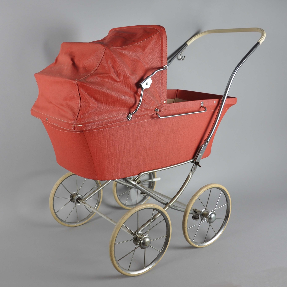 Dukkevogn med hjemmelaget madrass, dyne og pute. Vognen er av rød polyester med sammenleggbar kalesje. På sidene er det håndtak til å feste et løst vogndekke. Håndtak av kromt metall går ned på sidene av vognen, øverste del er lagt med hvit plast. Hjulene er hvite med eiker, ved høyre hjul er det en bremsespenne. Understellet på vognen er en lys treplate. På høyre håndtak er det satt på en refleks, av nyere tid.