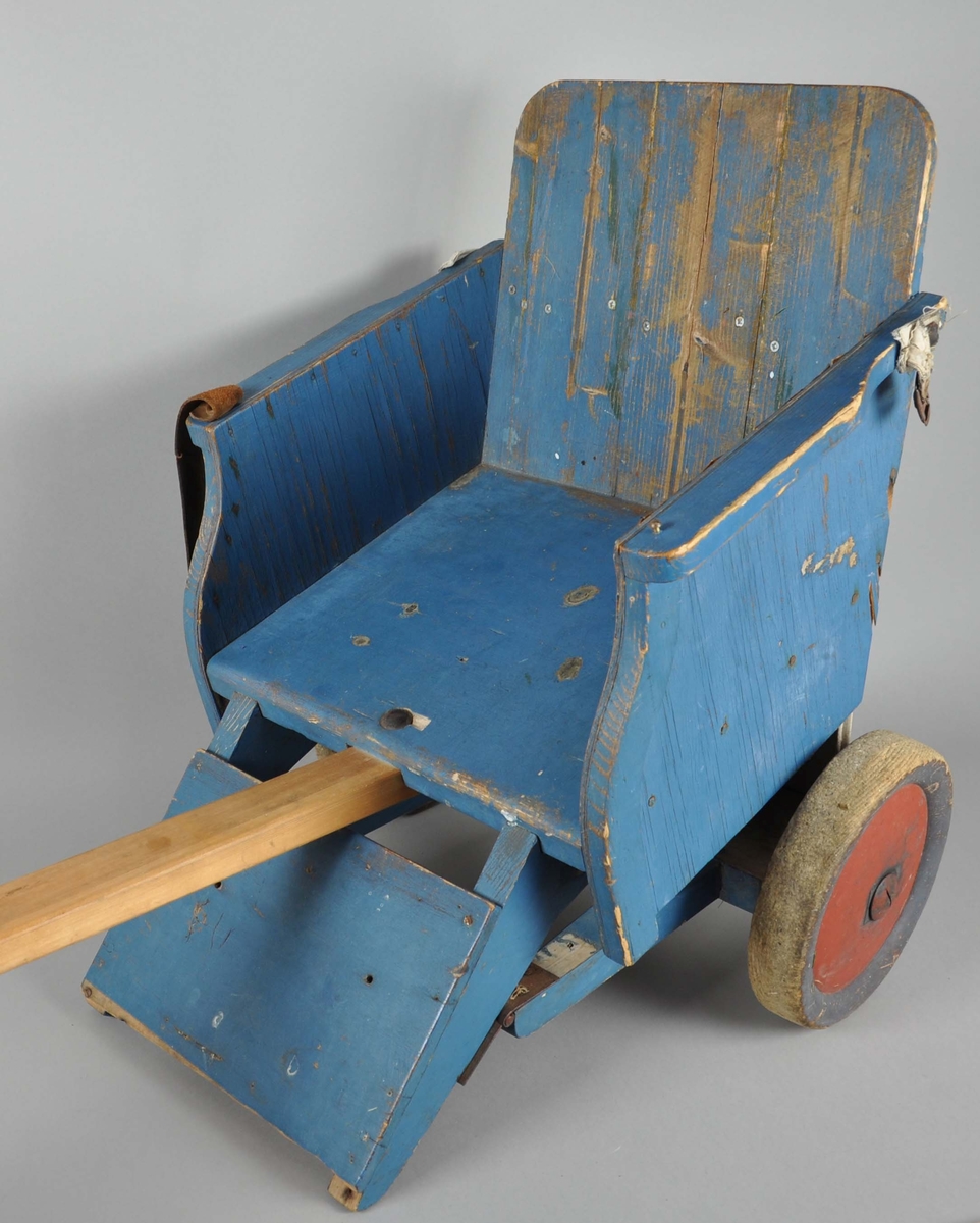 Håndlaget trilleav tre. Består av sete på to hjul, lærreim til belte, fotstøtte og draspak. Hjulene er av tre, og kapslene er malt røde. Setet står på to fjæringer fra madrass. Nederst på fotstøtten mangler det en oppstående list. Malingen er noe avslitt. Draspaken er trehvit og kan være erstattet i nyere tid.