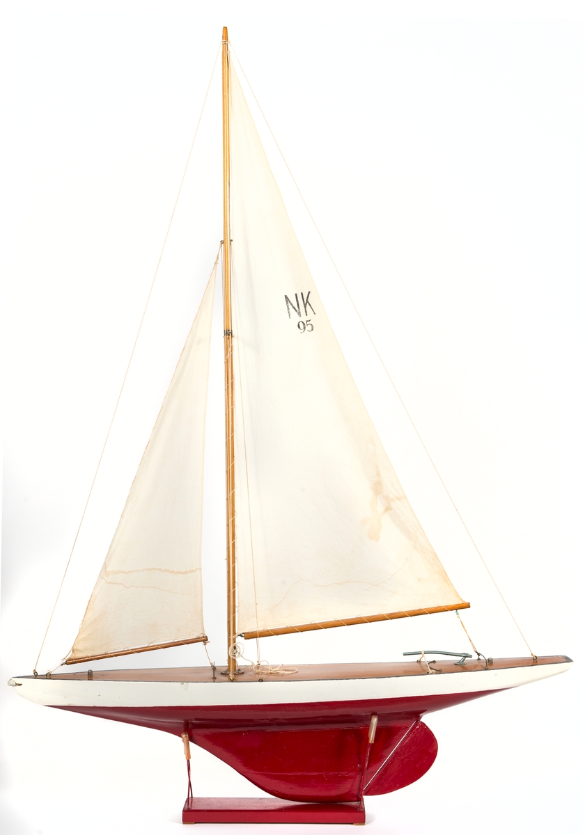 Modell av segelbåt, leksak. Tillverkad av NK senast 1935. 
Vitt skrov med röd bottenmålning. Fernissat nåtat däck och blyköl.
