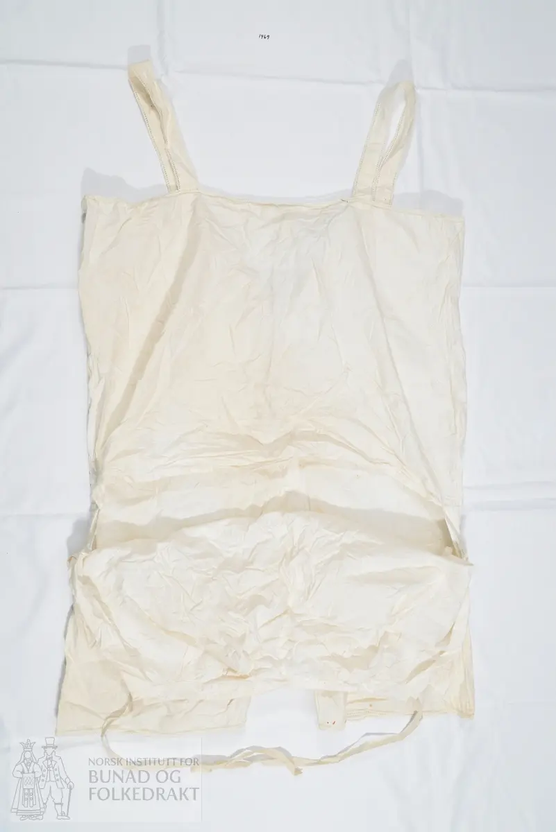 Chemilette, underbukse med overdel/ linnet, i hvit bomull. Knelang bukse, overdel med seler og åpning midt framme med knepping. Splitt i sidene og horisontal åpning bak. Overdelens ryggstykke stanser ved livlinjen. Lokk bak på buksen, med knytesnor øverst på lokket i hvert hjørne. Knytebånd til å knyte rundt livet. Pen avslutning på benene, med sydd hullfald og leggsøm, samt en smal heklekant helt ytterst.