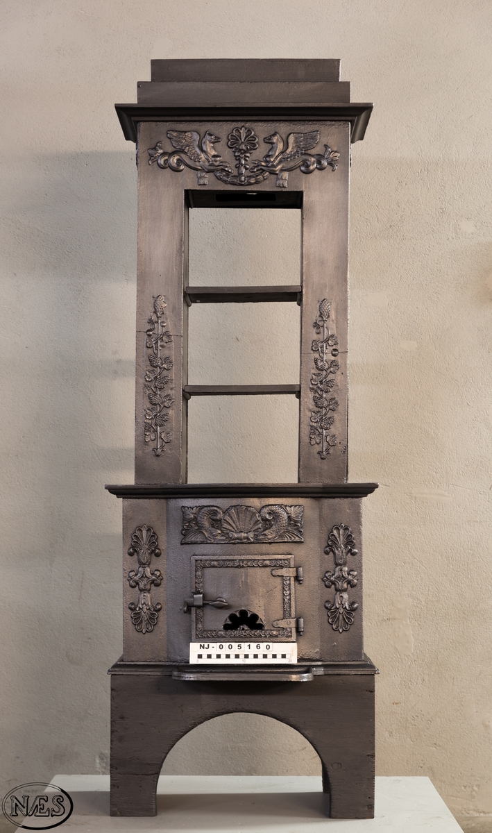 Ovnen består av to avdelinger. Underdelen har et frembuet midtparti med kamskjell flankert av sjødyr på hver side. På kortsidene skjold, løvehode og symmetriske ørner. På bakplate en krukke med symmetriske engler.
Overdelen er høy, firkantet med tredelt åpning. Empire ornamentikk og avtrappet topp. Øverst på frontplaten symetriske fabeldyr (pegasus) vendt mot hverandre.