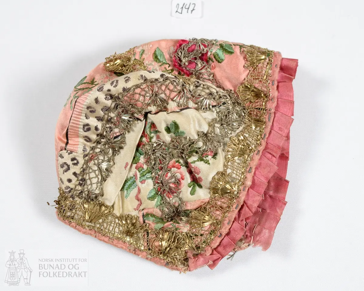 Dåpslue. Av rosa og hvit silkedamask med brosjert blomstermønster i to ulike rosa nyanser og grønt. Dekorert med metallkniplinger, i både sølv og gull. Smal rosa silkekanting rundt hele lua, i tillegg er et mørkerosa silkebånd i satengbinding foldelagt langs framkanten.  Foret med naturfarget linlerret. For og lue er vrangsydd, og så sydd sammen. Sømmene fra kniplingene er synlige på vrangen. Kun håndsøm. Stoffet er utnyttet med flere små skjøter.