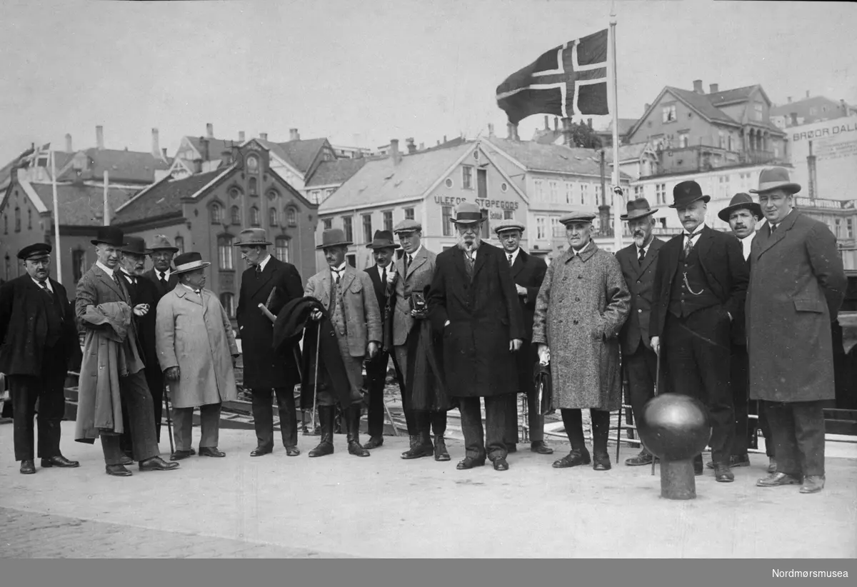 Gamle og nye herremoter på Piren i Kristiansund ca 1910. Aktverdige menn venter på viktig besøk. Tollbua bak, Flagg. Nordmøre museums fotosamling.