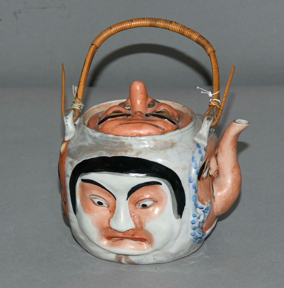 Rund kanne av keramikk med asiatisk dekor. Fire ansikter på korpus av kannen, lokket er også formet som ansikt. Tvinnet pinne fungerer som hengslet håndtak.