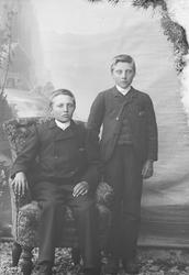 En unggutt  sitter i stol og ena annen står ved siden av.
N