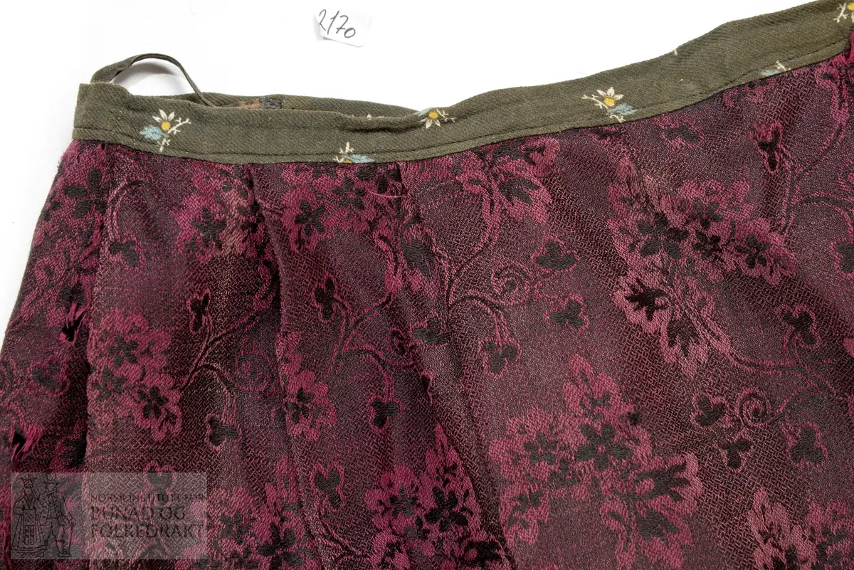 Brudestakk av mørk rosa og svart ulldamask. Av 3 høgder delvis sauma saman for hand og med maskin. Motfald midt framme, med eit breidt glatt parti midt framme. 3 breie legg på kvar side av motfalda. Midt bak eit 9 cm breidt rynka parti, med 2 breie legg på kvar side. 
Skonning og linning i trykt bomullskypert sauma på med maskin. Splitt midt bak lukkas med 1 par måla hetallhekter. I splitten møtes to jarekantar som er sauma ned med faldesting. På innsida av linninga er det påsauma to stoffhemper.
Stakken er bøtt i store stykkje både oppe ved linninga og i nedkant.