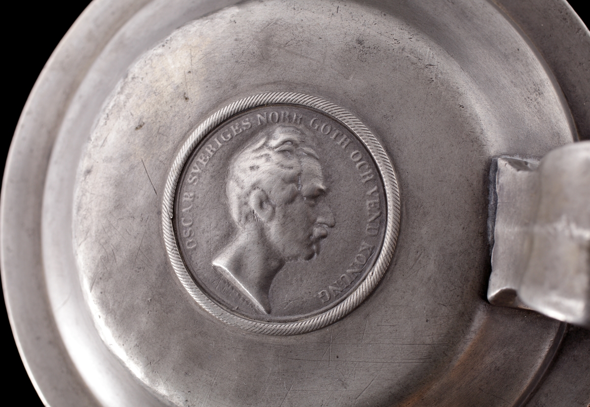 Stop i tenn, med lock. 
Cylindrisk modell med enkel banddekor. Locket försett med infälld medalj (mynt?) över Oscar I (reg. 1844-1859). 
Stämplar på undersidan av stopet.