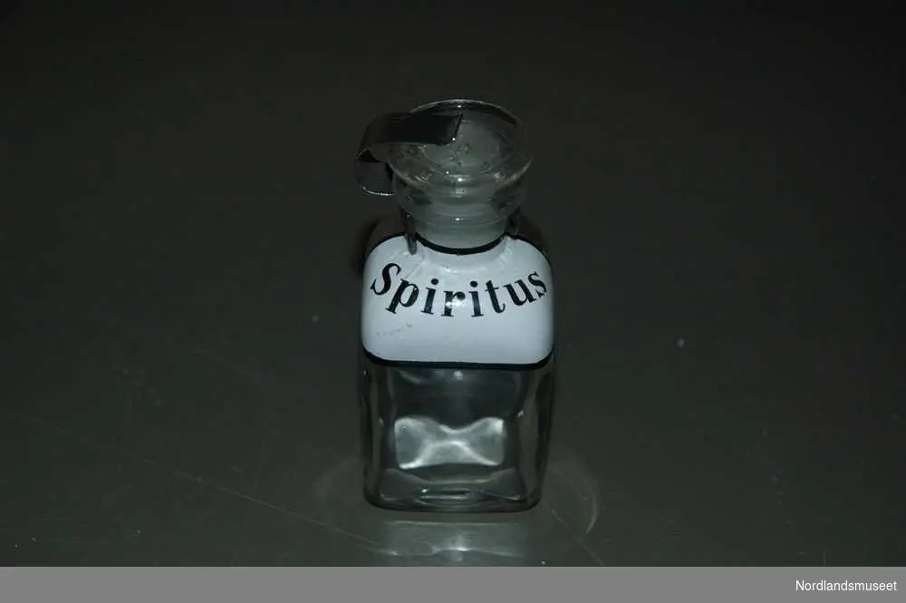 2 gjennomsiktige medisinflasker.
a: Den ene flasken står det "Spiritus" på. Den har en kork på toppen. Den har noe metall greier som holder den på plass.
b: Den andre flasken står det "Spiritus verd." Korken på toppen er feste med en streng rundt.