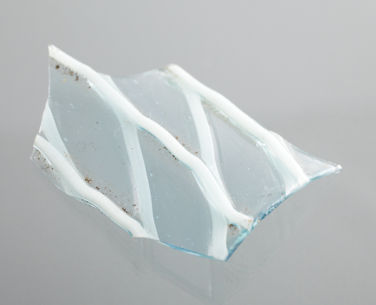 Två delar av ett ornerat venetianskt glas i svagt blå färg. På glaset finns två skikt pålagda vita trådar i ett grovt nätmönster (vetro a fili) i relief. Den större av skärvorna är en del av en mynningsrand i ett cylindriskt glas.