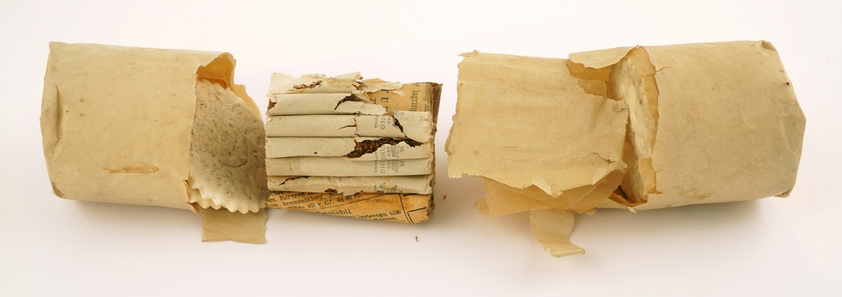 To deler av kjekspakke, i gul/grått papir, hvorav noe av papiret er blitt borte. En del av pakken, som skal ha vært midtdelen, består av sigarettene, innpakket i avispapir og snørt sammen med hyssing.