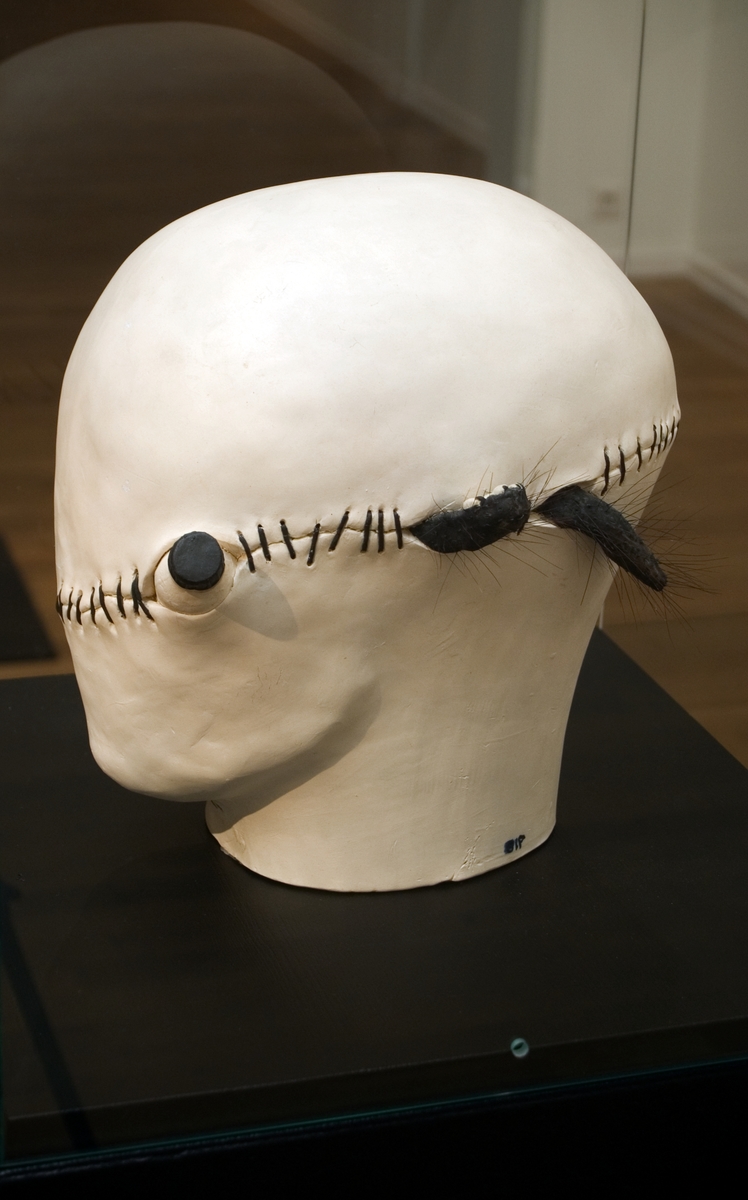 Keramisk skulptur av Britt-Ingrid Persson (BIP), "Rehabiliterad". En ljus huvudform, med en reva tvärs över ansiktet som "ihopsytts" med mörka stygn. Ur revan mellan stygnen sticker föremål fram; en kattass, en kattsvans och en liten del av en flaska med skruvkork. Konstnärens skulpturer i oglaserad lera från 1970-talet är politiska och i Rehabiliterad syns en kritik mot den tidens psykiatri, men inte utan en lite galet humoristisk vinkling.