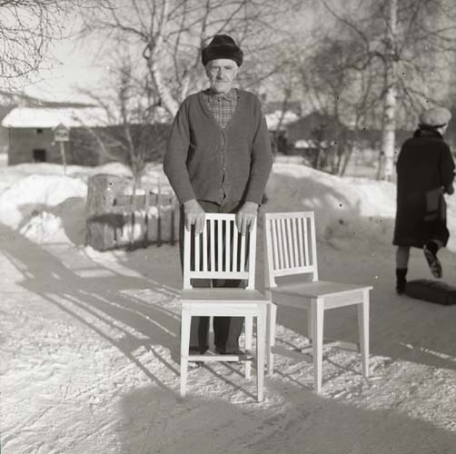 En man poserar i solen en vinterdag. Han står bakom två vita stolar.