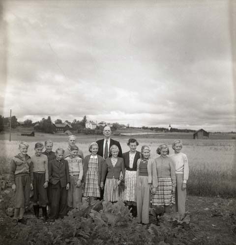 Några vuxna och ungdomar står uppställda på rad mitt i en gönsaksodling. I bakgrunden ser man en gård, en kyrka och en lada. Unga Odlare, Söderhjelm med, augusti - september 1950.