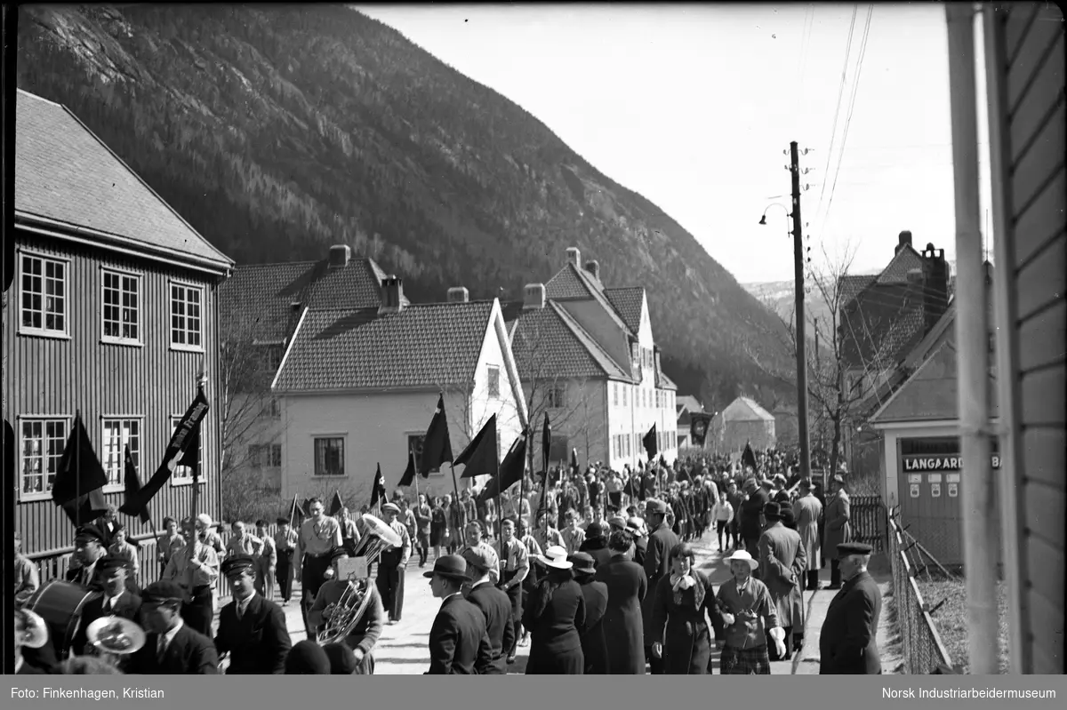 Rjukan Framlag i 1. mai-tog i Sam Eydes gate. Korps spiller i toget. Øvre og nedre Sing Sing sees i bakgrunnen, samt blåkiosken med skiltet "Langaards" på veggen.