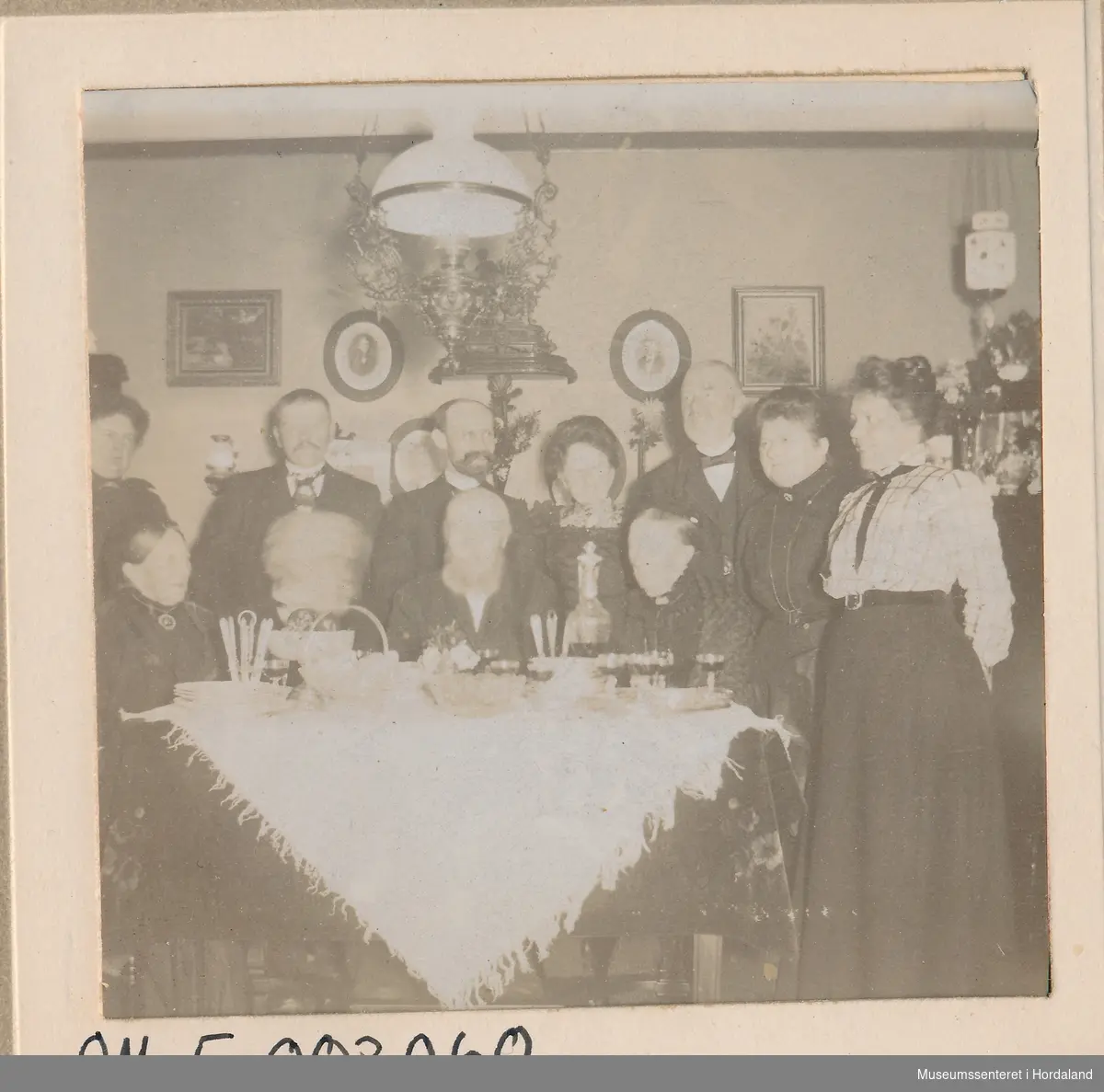 amatørfotografi av sju kvinner og fire menn i finkle bak eit dekka bord med kvit duk, karafflar og glas i ei stove med bilder på veggane og glas/messing taklampe over bordet
