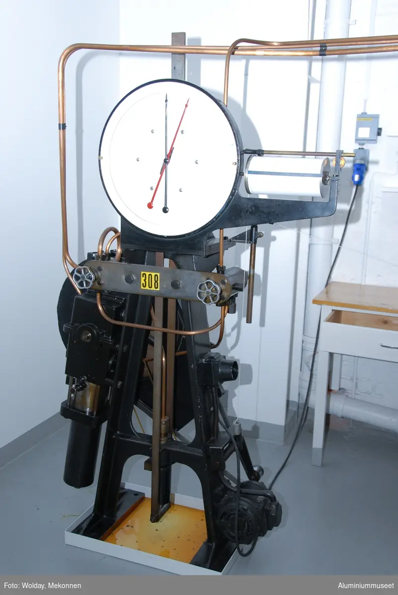Strekkprøvemaskin for metaller.  Leverandør: Alfred J. Amsler & Co. Schweiz.  Datering: ca. 1925-1966. Teknikk: Hovedenhetene er støpt i stål og delvis maskinert.  Enkelte deler er fremstilt av valset stål og hydrauliske overføringer er kobber-rør.
Form: To stk. maskinenheter.  En betjeningsenhet og en strekkenhet som er forbundet med hydrauliske rør.
Tilstand: Maskinen er operativ, men har hydrauliske lekasjer.
Utfyllende opplysninger:
Betjeningsenheten består av en hydraulisk pumpe som via 2 stk. reguleringsventiler leverer olje til sylinderen for pendelmanometeret og til hovedsylinderen på toppen av strekkenheten.  Strekkprøvene plasseres i klemmebakkene på strekkenheten, blyanten settes på papirrullen og høyre kran på betjeningsenheten åpnes.  Etterhvert som trykket økes blir prøven strukket helt til brudd.  Under strekket slår pendelen ut tilsvarende trykket i hovedsylinderen og strekk-kraften kan til enhver tid avleses på skala-skiven.  Via et snorsystem fra overbjelken hvor øvre klemmbakke sitter, vil strekkforløpet bli avtegnet på en papir-rull på betjeningsenheten og strekkforløpet blir avtegnet slik at hele strekkforløpet og flytgrensen kan avleses.  Ved bruk avstenges hovedkranen umiddelbart og da vil den sorte pila umiddelbart gå tilbake til null-posisjon., men den røde pila blir stående igjen og viser bruddkraften.  Med den venstre kranen slippes hovedsylinderen ned til utgangsposisjon igjen.  Tykkelse og bredde på prøvestaven blir målt før prøvetaking og staven blir også risset med tynne paralelle strekker med 10 mm mellomrom.  Se risseapparat HAM.79.  Etter brudd blir prøvestaven satt sammen igjen og målt over en lengde på 5 riss med bruddet ca. midt i mellom. Den nye lengden i forhold til 50 mm viser forlengensen som blir utregnet i prosent.  Bruddfastheten er avlest bruddkraft delt på tverrsnitt.  Flytegrensen avleses av det tegende diagrammet på papir-rullen pluss 0,2%.  Maskinen kan maks. yte 10 tonn, men pga. unøyaktig avlesing ved tynne prøver blir pendelmanometeret innstilt ettter forventet bruddstyrke.  Maskinen har 4 innstillnger: Maks.: 1000 kg: Nest nederste hull. Uten lodd. Maks.: 2000 kg: Nest øverste hull. Uten lodd. Maks.: 5000 kg: Nederste hull.  Med Lodd. Maks.: 10000 kg.: Nest øverste hull. Med lodd.
Maskinen står i dag 08.01.2007 i psosisjon, 5000 kg.  Maskinen kan også brukes til trykkprøving, f.eks. sveiseprøver, ved å plassere prøven på rullebakkene oppe på strekk-bommen.  22.01.2008 Kjell Grette.