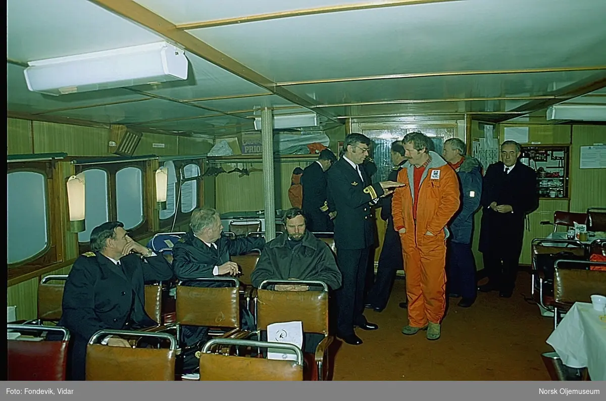 Bilder fra salongen til et skip. Det er flere menn samlet i salongen, hvorav flere av dem har uniformer fra Den kongelige norske marinen, og noen har arbeidsklær med logoen til "NUTEC" på.
Bildene kan muligens være tatt ombord fartøyet M/S "Halsnøy", bygd i 1957, for HSD