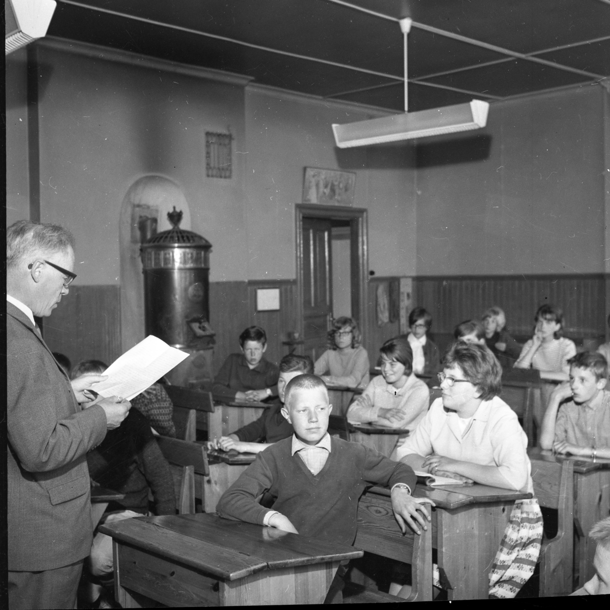 Sista lektionen i Uppgränna skola. Rektor Erland Skoog står framför eleverna, som sitter i sina bänkar, och läser i ett papper. I mitten sitter Maud Enehed, snett hitom henne Ros-Marie Jonsson med glasögon och pojken till höger är Peter Weidner.