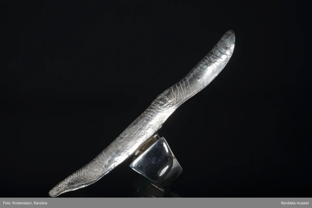 Ring av silver, överdel i form av avlång avgjutning av fingerrygg. Underdel bestående bred ring med slits på ena sidan. Undertill graverad "Cornelia".
/Leif Wallin 2018-02-09