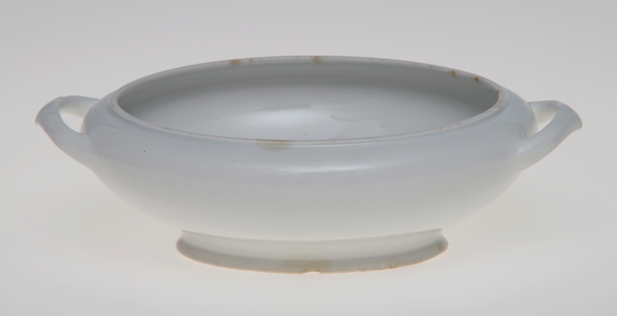En oval serveringsbolle, muligens en liten terrin, som mangler lokk. Den er laget av steintøy og er hvitglasert. I hver ende er det en enkel hank med enkel innstøpt dekor.