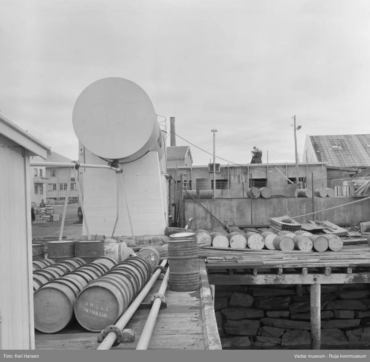 Vadsø sentrum 1959. Shells tankanlegg for drivstoff under bygging. Tønner og bygningsmateriale lagret på kaia. Til venstre i bildet ser vi litt av  H.F.Esbensens forretningsbygg og deler av Telegrafen. Midt i bildet skimtes taket til Meieriet og pipa.