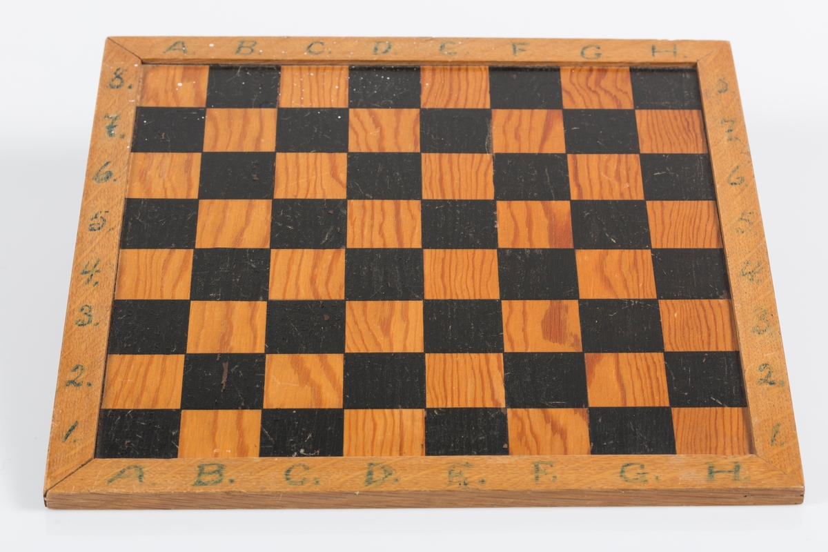 Kvadratisk sjakkbrett av lakkert tre med treramme. Sjakkfeltene er trefarget og malt sort. Bokstaver og tall skrevet med blågrønn farge.