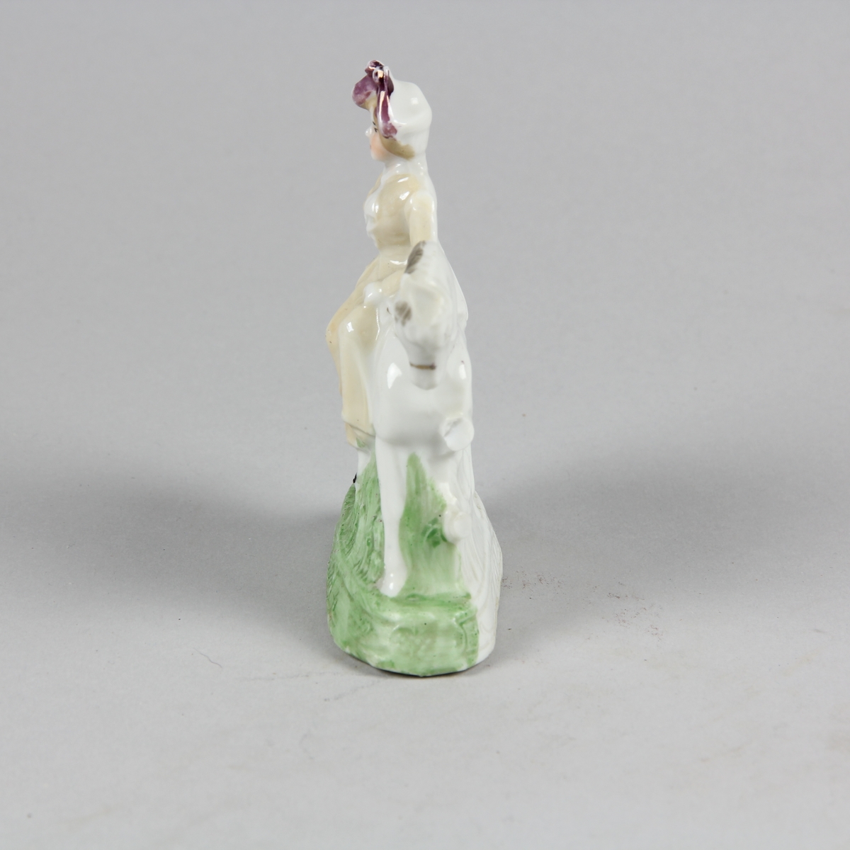 Prydnadsföremål av vitt porslin i form av liten skulptur föreställande en kvinna som sitter på en häst i damsadel. Kvinnan är klädd i lång gulvit klädnad och lila hatt. Hästen vit med svart man. Grönt gräs och grön sockel. Endast framsidan, vänd mot betraktaren är bemålad.