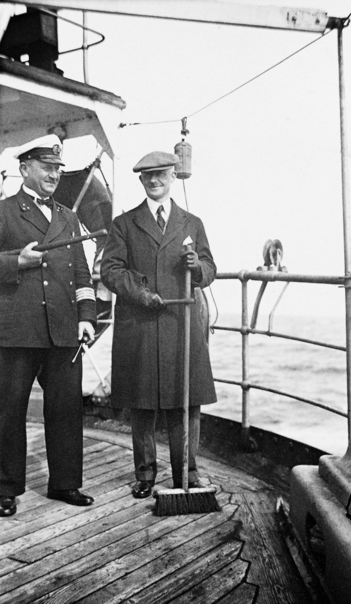 Fra akterdekket på en dampbåt, antakelig i 1930-åra.  Til venstre ser vi en uniformert mann, sannsynligvis fartøyets kaptein, og til høyre en mann som var kledd i frakk og hadde sixpencelue på hodet.  Sistnevnte holdt en feiekost i handa, men var åpenbart passasjer, ikke et besetningsmedlem på båten.  Fotografiet er fra albumet etter Egil Abrahamsen, som fra 1928 arbeidet for den engelske banken Wm Brandt’s Sons & Co.  Båtpassasjeren på fotografiet er muligens forretningsmannen «Alec», som Abrahamsen sommeren 1936 ledsaget på en tur til Arkhangelsk, der Abrahamsen tidligere hadde arbeidet for nordmenn som hadde investert i russisk trelastindustri.  I Abrahamsens memoarbok – «Vinter i Seregovoskogen – Erindringer fra det norske trelasteventyret i Russland» (2015) beskrives turen til Arkhangelsk med «Alec» slik:

«Et annet år kom jeg en tur til Russland igjen, det var sommeren 1936.  En fremstående importør fra Hull som vi kalte for Alec, ville en tur til Arkhangelsk for å se på en del trelast og ba meg ordne reisen og bli med ham som tolk.  En av mine sjefer var også med for personlig å se seg om i Russland.  Alec var, selv til engelskmann å være, en usedvanlig rolig og behersket kar.  Vi to en båt til Leningrad, og ble sendt videre til Moskva, hvor vi bodde på Metropol noen dager og beså oss.  Akkurat da kom den sagnomsuste Stalins konstitusjon av 1936, og vakte voldsomme diskusjoner og begeistring blant russerne.  Konstitusjonen inneholdt mange pene punkt om organisasjonsfrihet, talefrihet o.s.v., og folket trodde at endelig, gir Fader Stalin oss friheten tilbake som vi tilkjempet oss i 1917.  Hva ble så følgene av at så mange begynte å uttale seg så høyt og fryktløst?  Jo, Stalins hemmelige politi fikk satt opp nye lister over folk som var deres motstandere, store og små partifolk og andre, og disse ble i tusenvis henrettet under de påfølgende prosessene i 1937 og 1938.  Var det hensikten med konstitusjonen?

Omsider kom vi til Arkhangelsk, men der møtte vi en skuffelse.  Det eneste skikkelige hotellet var brent ned og vi ble innkvartert på et lite, elendig, overfylt et.  Nå var Alec av den type som helt fra barndommen av var vant med et kaldt bad om morgenen og et varmt om kvelden.  Hvis han ikke fikk dette, ble han i dårlig humør.  Nå hadde jeg hørt at det var et eneste pent rom med bad på hotellet, men det var opptatt.  Jeg fikk derfor til hotellbestyreren og spurte om han kunne ordne det slik med vedkommende gjest at Alec kunne få ta sine to bad hos ham.  Men det viste seg ikke å være mulig.  Værelset var nemlig besatt av en marskalk Eidemann av Den røde armé, og «han er en så stor mann at jeg tør ikke spørre ham engang», sa bestyreren.

Alec ble i så dårlig humør at vi reiste snart hjem.  Et par måneder senere satt jeg på kontoret og leste i Times om henrettelsen av fem russiske marskalker.  Nettopp da kom Alec inn for å snakke med meg.  Jeg sa: «Husker du marskalk Eidemann i Arkhangelsk?»  «Den fyren med badet», sa Alec.  Jeg nikket bekreftende og sa at nå var han skutt av Stalin.  Han kunne ikke låne oss badet, da ingen torde be ham om dette, og nå ble han skutt av Stalin på en anklage for å være spion for Tyskland og Japan.  Denne anklagen var falskelig fabrikkert av Stalin og i dag, 20 år senere, er Eidemann og de andre henrettede marskalkene rehabilitert av Khrustsjovs regjering.
På reisen med Alec i 1936 fikk jeg anledning til å besøke en stor «selo» (landsby) ved Dvina.  Jeg hadde alltid ønsket å se denne landsbyen; skjønt jeg mange ganger passerte den på elven, enten med passasjerbåt eller bukserbåt hadde det ikke vært mulig å stoppe der.

Men en dag overlot et trelastkontor som Alec og jeg hadde noe å gjøre med, oss en fin motorbåt med tre manns besetning for å ta en sightseeing-tur oppover elven Dvina.  Til min glede tok karene oss så langt som til en stor, meget berømt landsby, Kholmogory, ca. 80 km. syd for Arkhangelsk, og der hørte vi og så vi mange interessante historiske ting.  En del av disse hadde jeg riktignok hørt om allerede den første vinteren (1908/09) jeg tilbragte i Privodino, idet det på den tid ble fortalt alle skolebarn i hele Russland om en begavet bondegutt fra Kholmogory, som i 1730 hadde gått til fots helt til Moskva for å søke seg lærdom, og som ble Russlands største sønn som videnskapsmann, poet og sprogforsker.  Jeg kan ennå huske uttrykket til de små landsbybarna som jeg satt sammen med, når de hørte lærerens fascinerende fortellinger om Mikhail Lomonosov, 19-åringen fra Kholmogory.  Russlands største sønn – en bondegutt!»