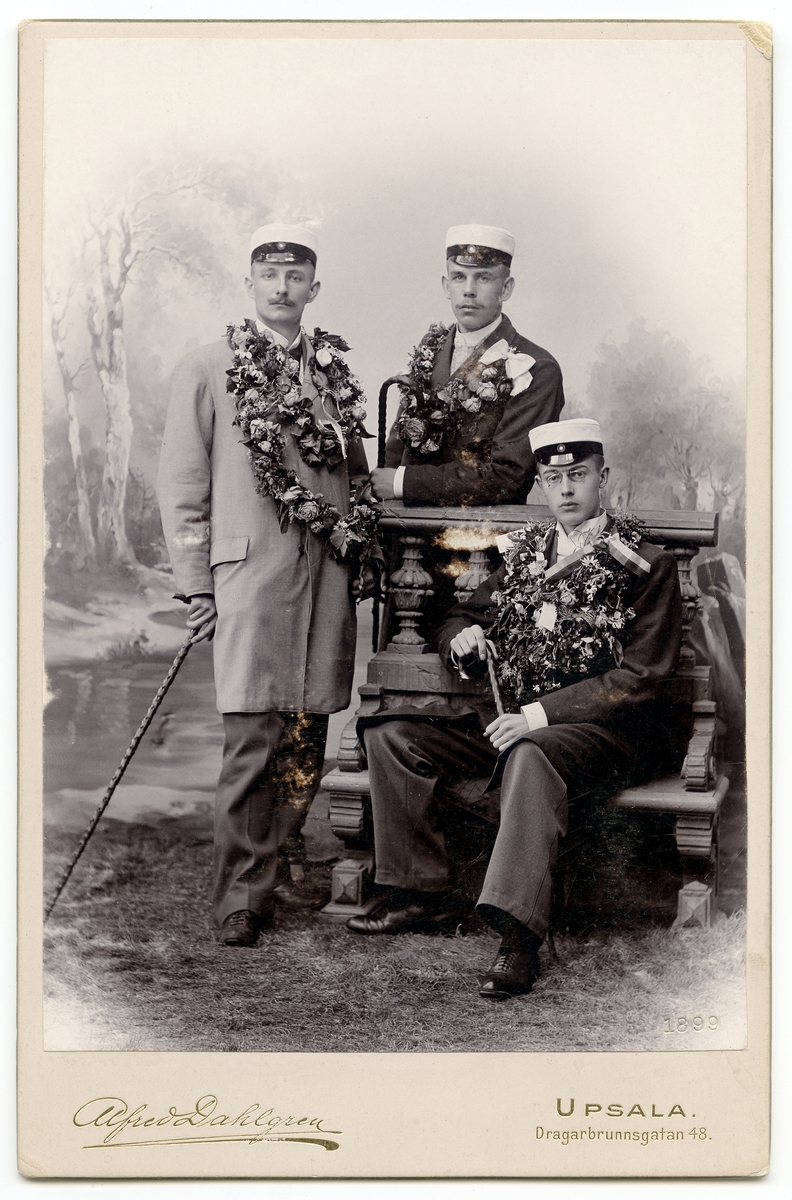 Kabinettsfotografi - tre män iklädda studentmössor, med käpp och garnerade med blommor. Den ena sitter på bänk, övriga två står, Uppsala 1899
