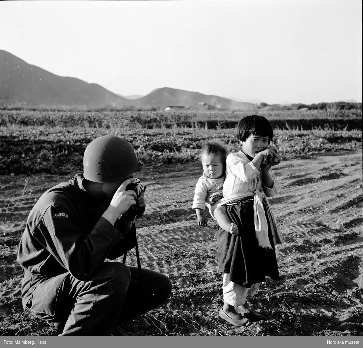 Koreakriget. En svensk pressfotograf tar en bild på en liten flicka som bär ett spädbarn på ryggen.