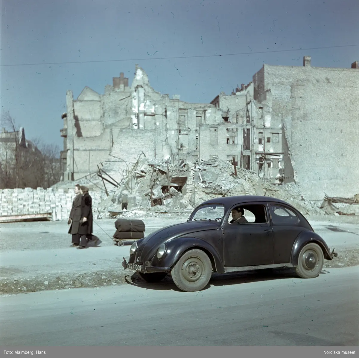 Berlin. En soldat ur de brittiska ockupationstrupperna kör en Volkswagen. I bakgrunden husruiner och två kvinnor med en kärra.