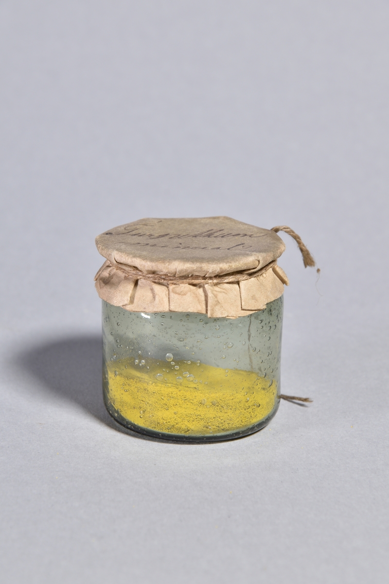 Burk av klart glas, cylindrisk med överbundet lock av papper, fäst med tunt snöre. Innehåller gulbrunt pulver.