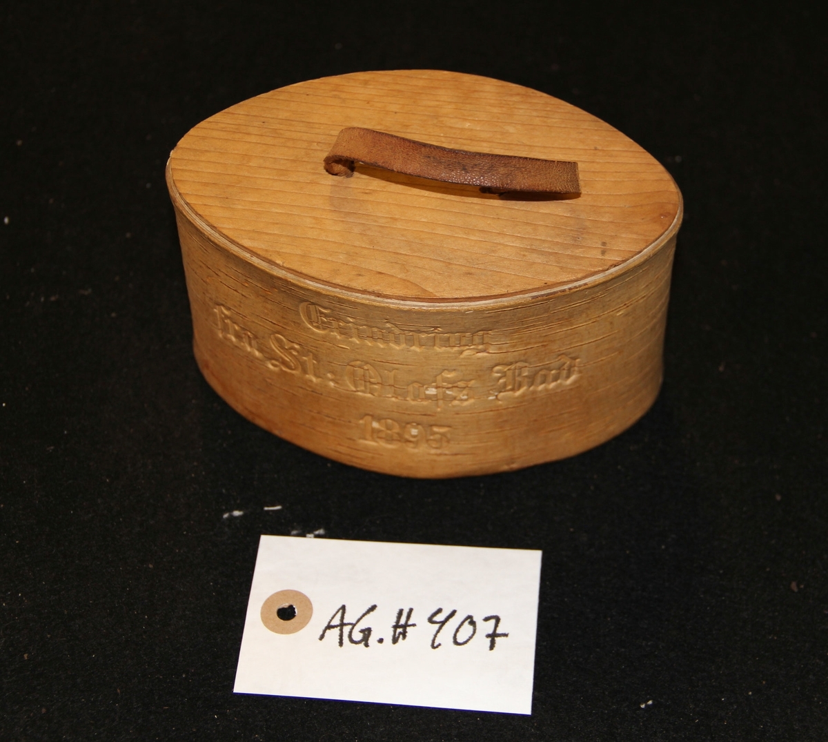 Oval tretine  og lokk med lærhåndtak. Inskribsjon på framsiden av tinen