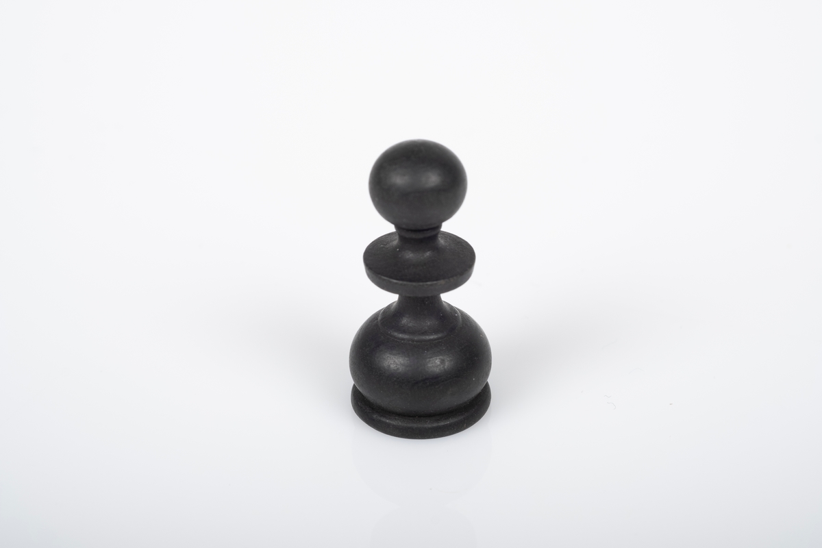 Sjakkbrikke (bonde) av tre. Brikken er dreid og malt/lakkert svart.