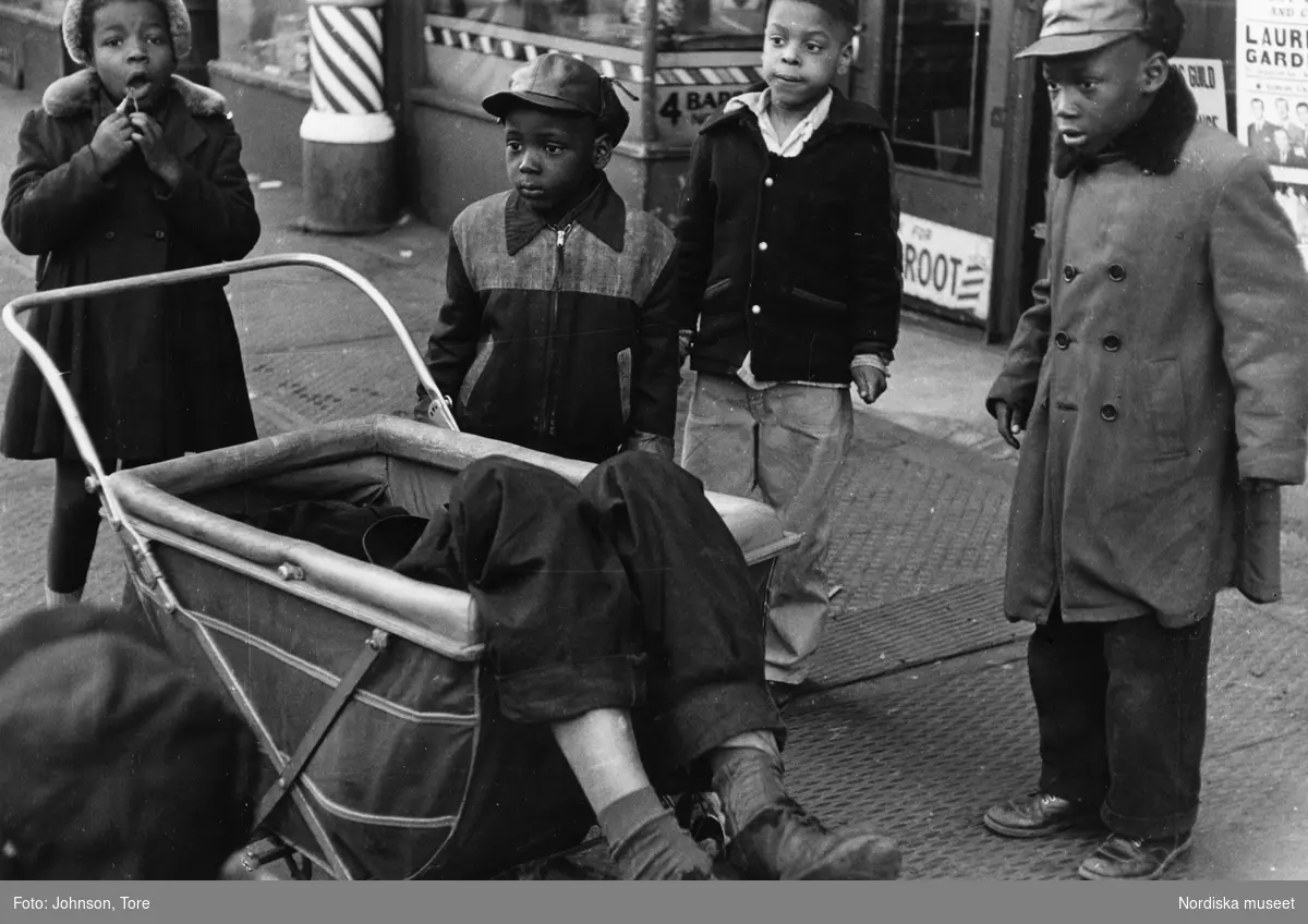 En grupp barn bredvid en barnvagn i vilken en utslagen man ligger. Lenox Avenue, Harlem, New York, USA.