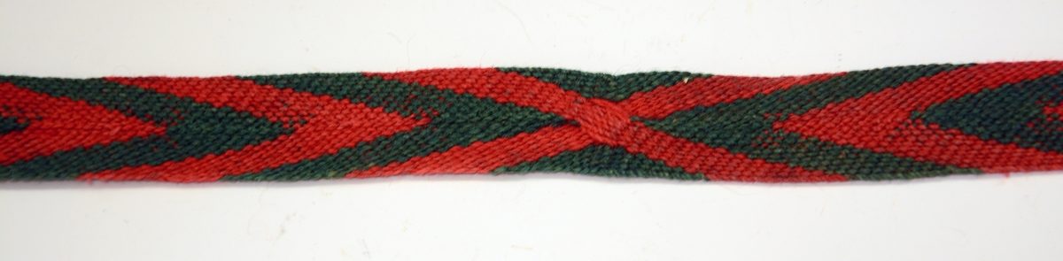 2 stk. strømpebånd i fingerfletting. Fiskebeinmønster i rødt og grønt. Renningen er formet til en dusk i hver ende.
