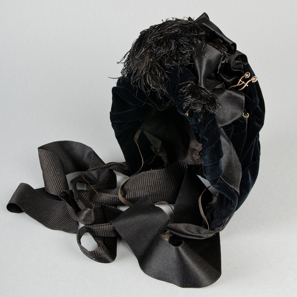 Av svart sammet, rynkad och draperad på ståltrådsställning. Svarta sidenband och strutsfjädrar samt tre hattnålar. Inuti i ett firmamärke av papper Lina Lundberg, Modehandel, Eskilstuna. 

