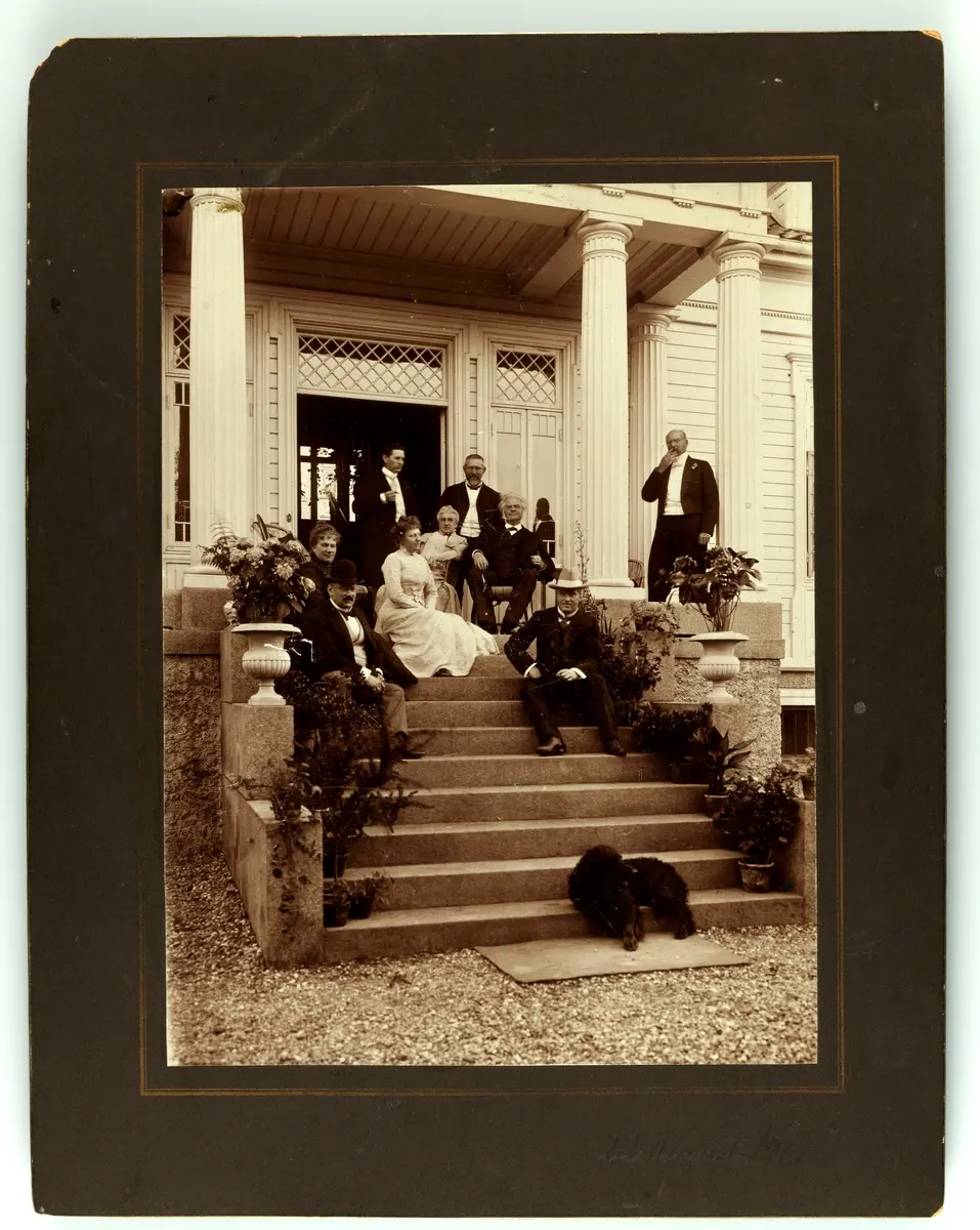 Bjørnstjerne og Karoline Bjørnson sitter omringet av venner og familie (og en hund) på en trapp i sommerlige omgivelser.

Fra venstre: Agnes Gude, Hans Aimar Mow Grønvold (foran), Gina Oselio, uidentifisert mann (stående bak), Karoline Bjørnson, Gustav Storm, Bjørnstjerne Bjørnson, Bjørn Bjørnson og Erik Anker Gude.

22. august 1901, Odden, Bygdøy